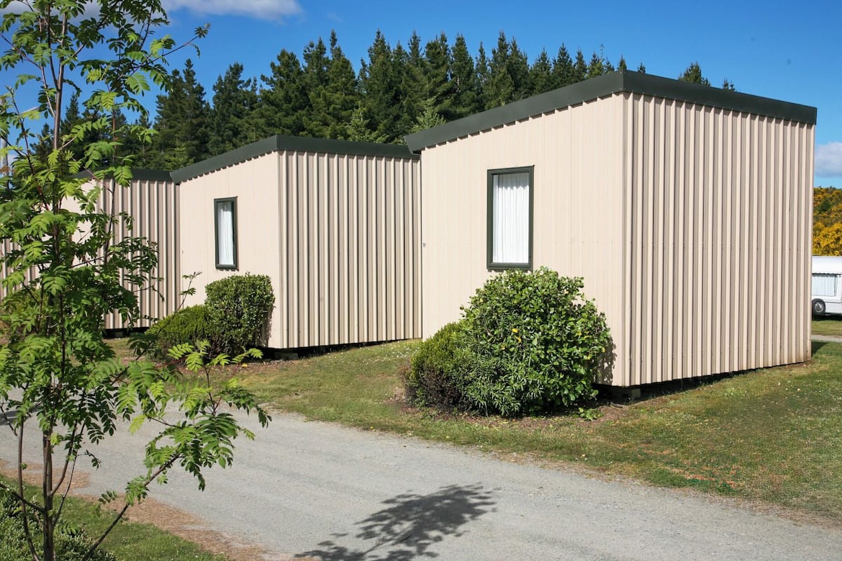 A row of two-story mobile homes with a flat roof, each featuring a window and surrounded by small bushes.