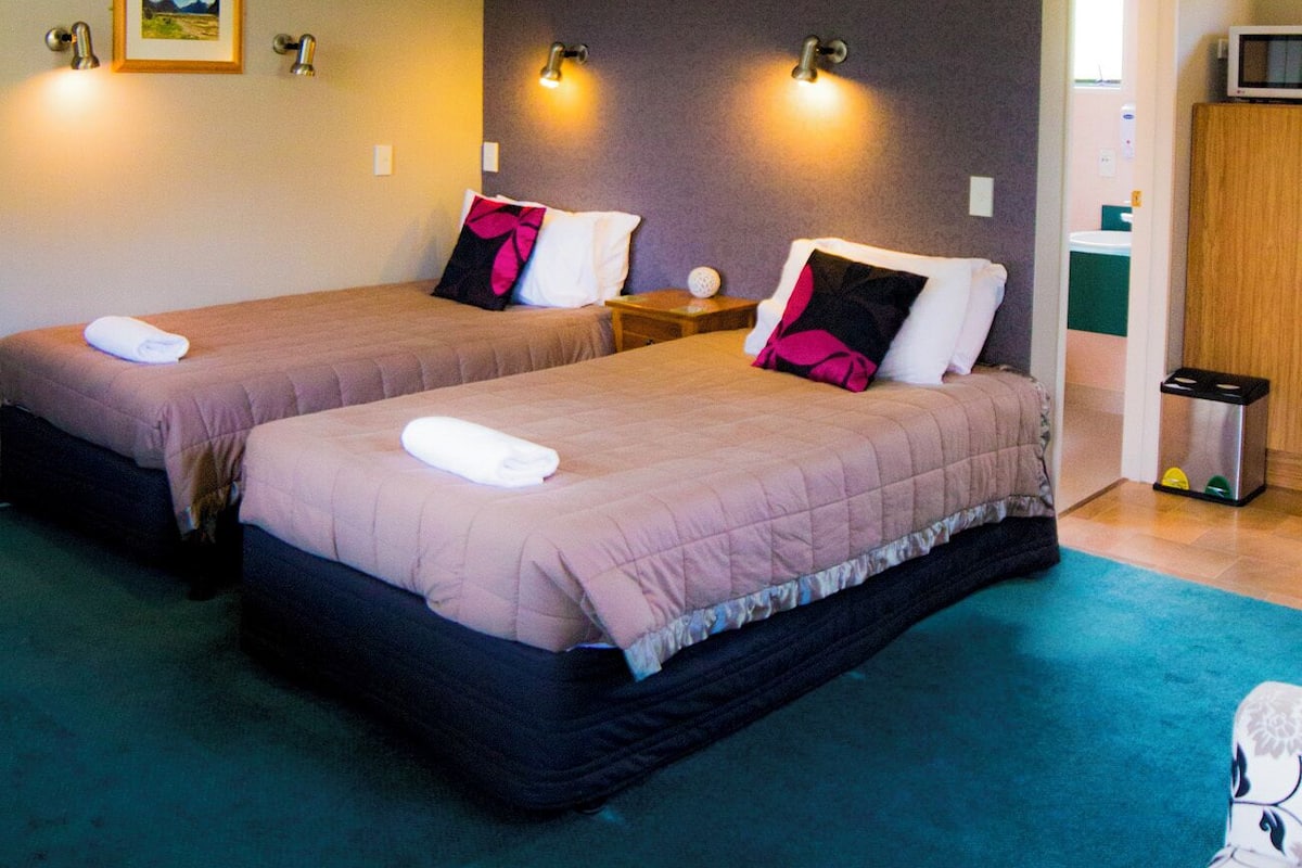 A room with two beds, each with a pillow and a blanket, a small table with a lamp, and a wall-mounted clock.
