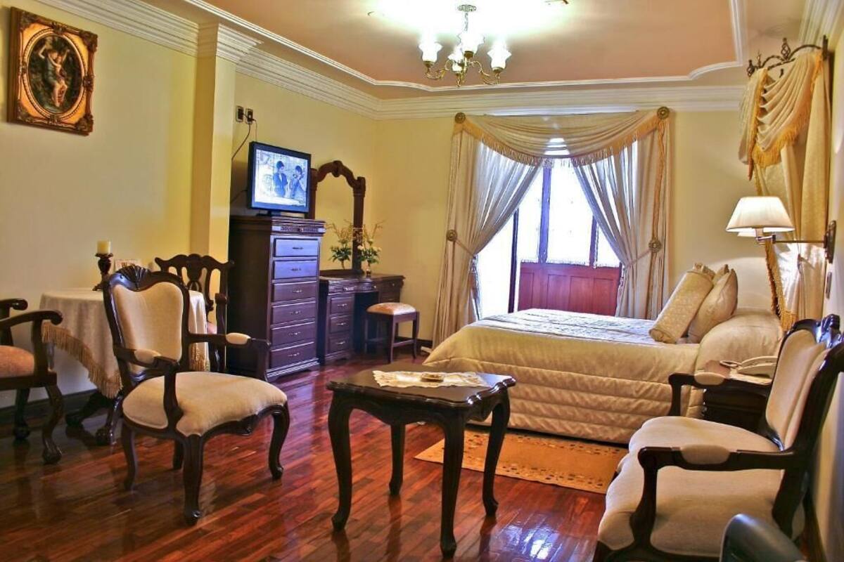 Una habitación con una cama, dos sillones, un aparador de madera, un televisor y una araña.