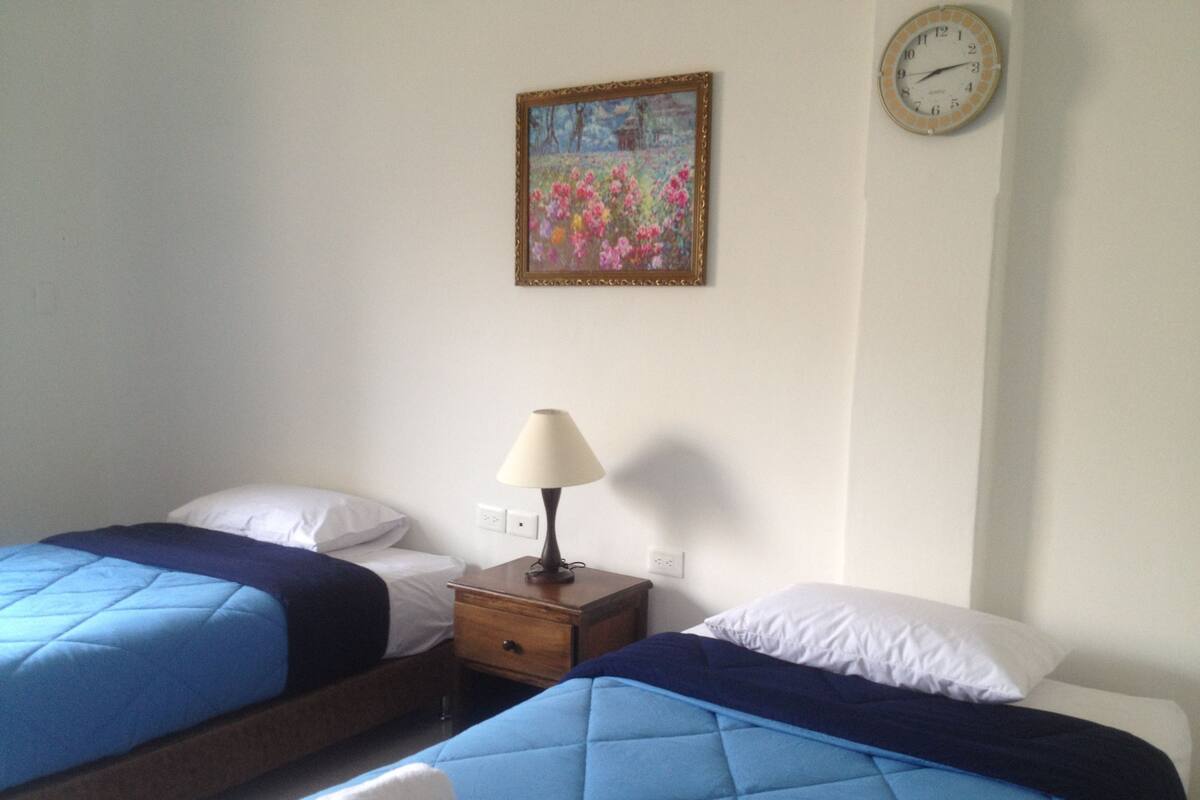 A hotel room with two beds, a nightstand, a lamp, a clock, and a painting on the wall.