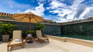 Un área junto a la piscina con dos tumbonas, una sombrilla grande y un muro de piedra.