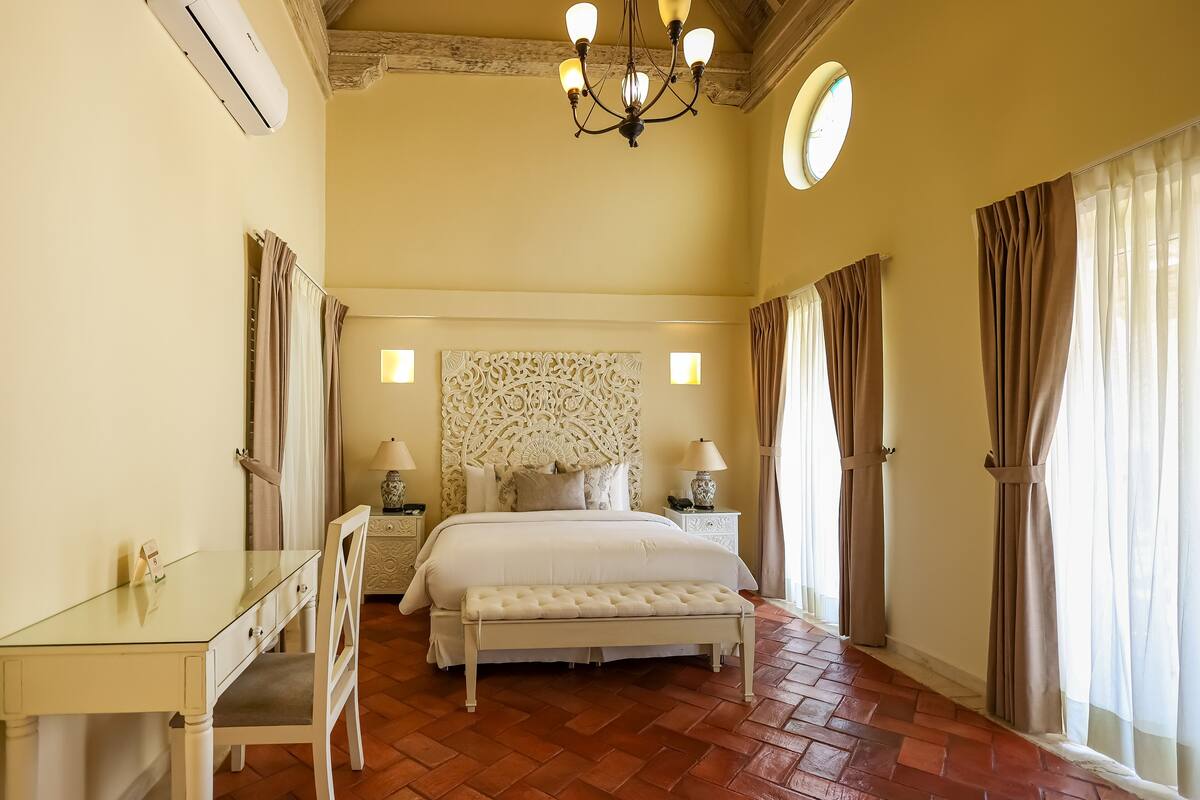 Une chambre à coucher comprenant un lit, des tables de chevet, un bureau, une chaise, un banc, un lustre et des rideaux.