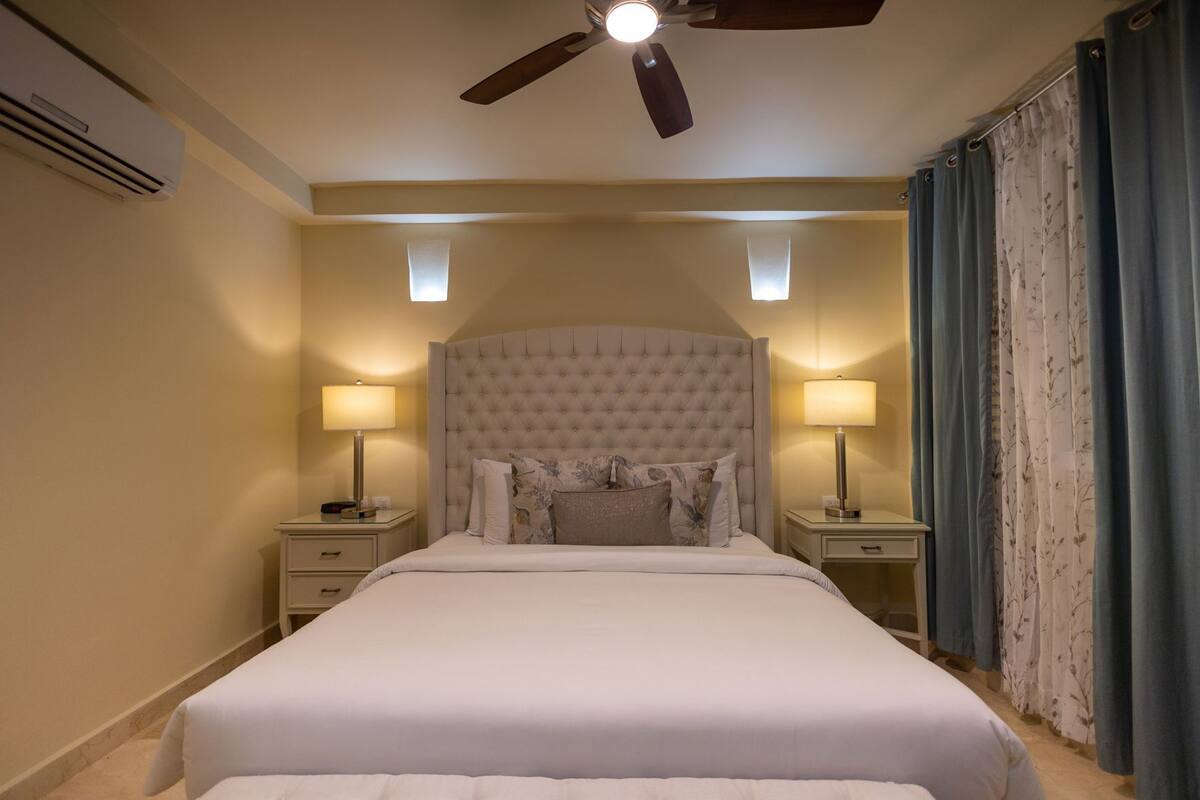 Une chambre à coucher avec un grand lit, deux tables de chevet, un ventilateur de plafond et une fenêtre avec des rideaux.