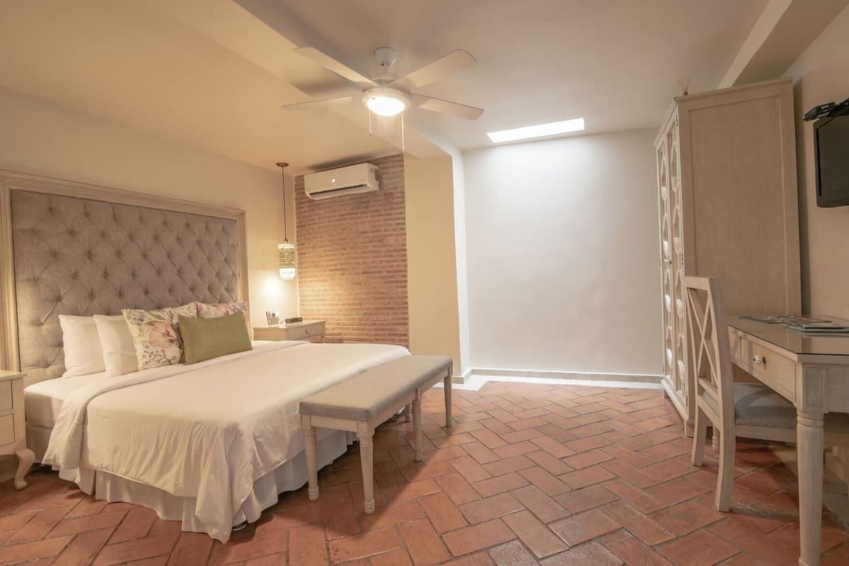 Une chambre à coucher comprenant un lit, un banc, un bureau, un ventilateur de plafond, un appareil de climatisation et un mur en briques.