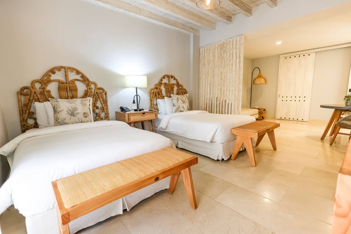 Une chambre d’hôtel avec deux lits, un banc en bois, une table de chevet avec une lampe et une petite table avec une chaise.