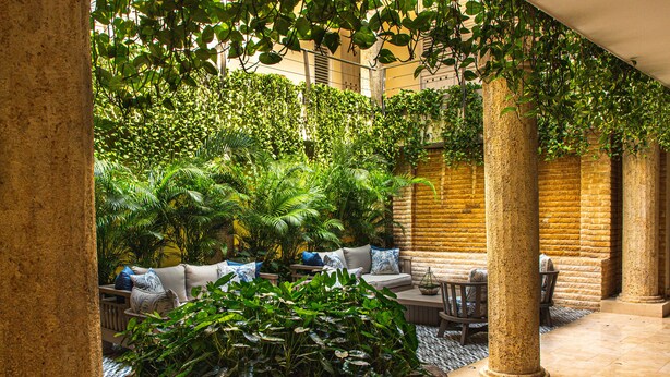 Un espacio de estar al aire libre cubierto, con pared de ladrillo, columnas y abundante vegetación.