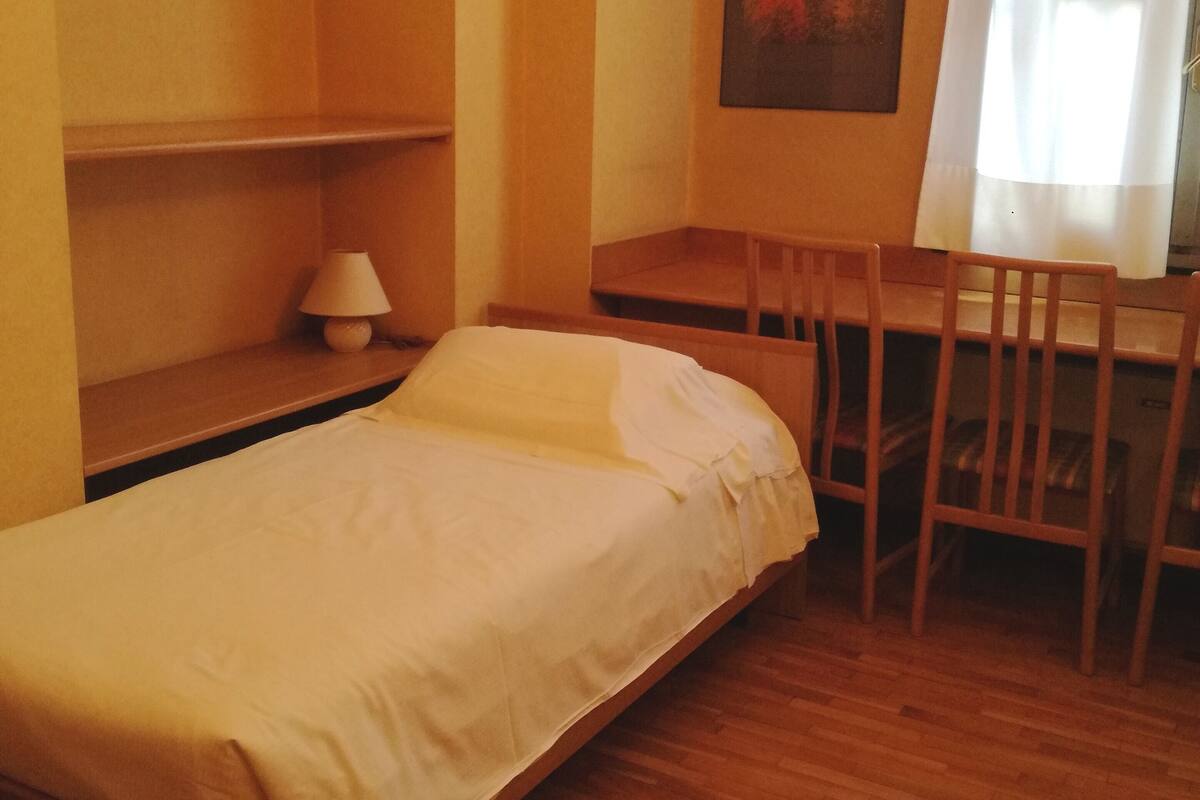 Una cama individual con edredón blanco, un escritorio de madera, una silla, una lámpara y un cuadro en la pared.