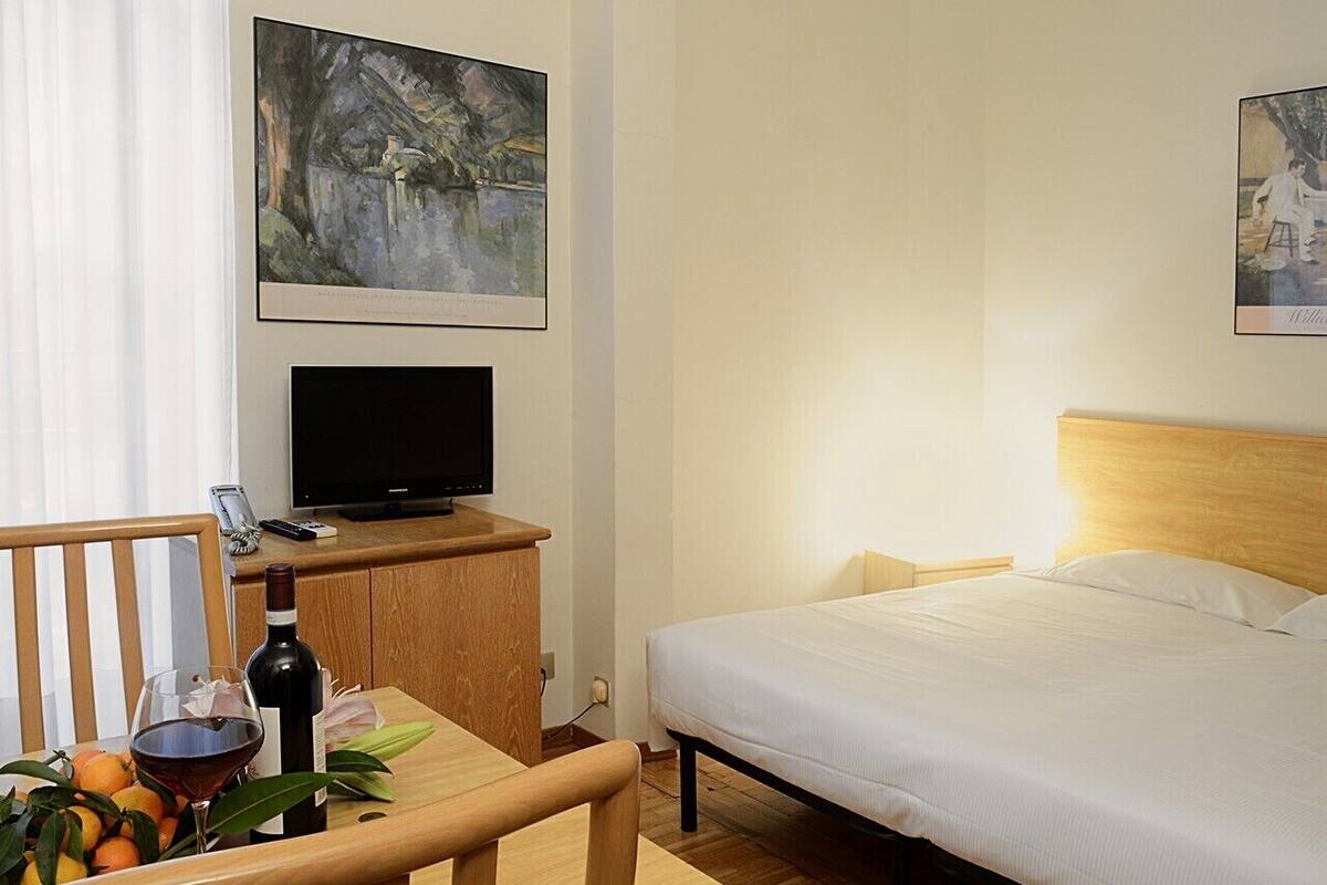 Una habitación de hotel con una cama, un escritorio con televisor, una silla, una mesa con una botella de vino y naranjas, y dos cuadros en la pared.
