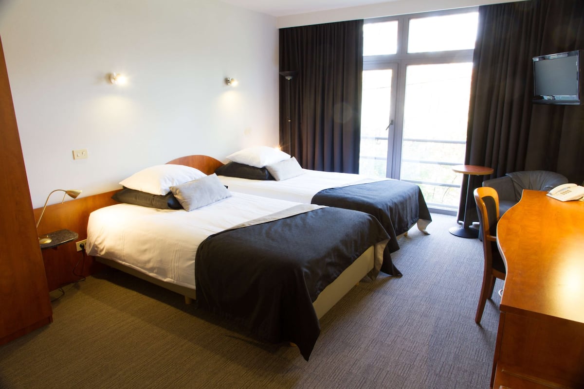 Quarto de hotel com duas camas, uma escrivaninha, uma cadeira e uma televisão na parede.