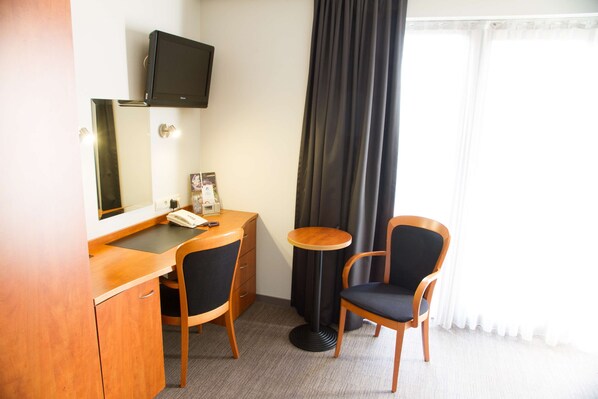 Habitación de hotel con un escritorio de madera, dos sillas, un televisor montado en la pared, una mesita redonda y una ventana con cortinas.