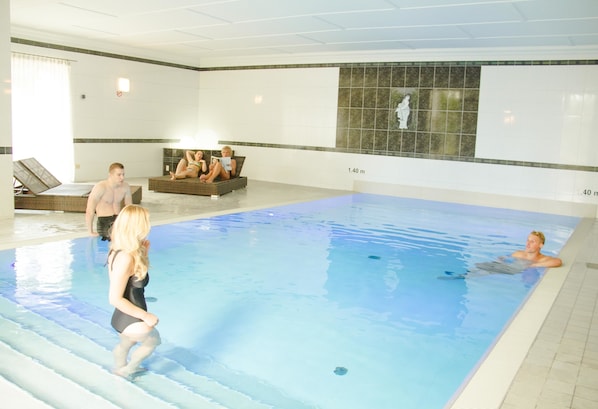 An indoor swimming pool with clear water, a tiled wall with a mosaic, and a lounge area with sunbeds.