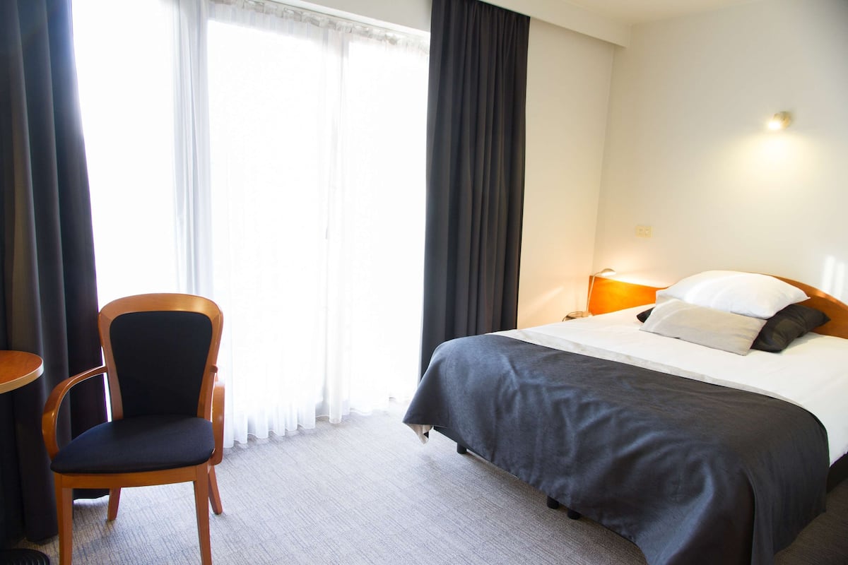 Quarto de hotel com cama, cadeira, mesa de cabeceira e janela com cortinas.
