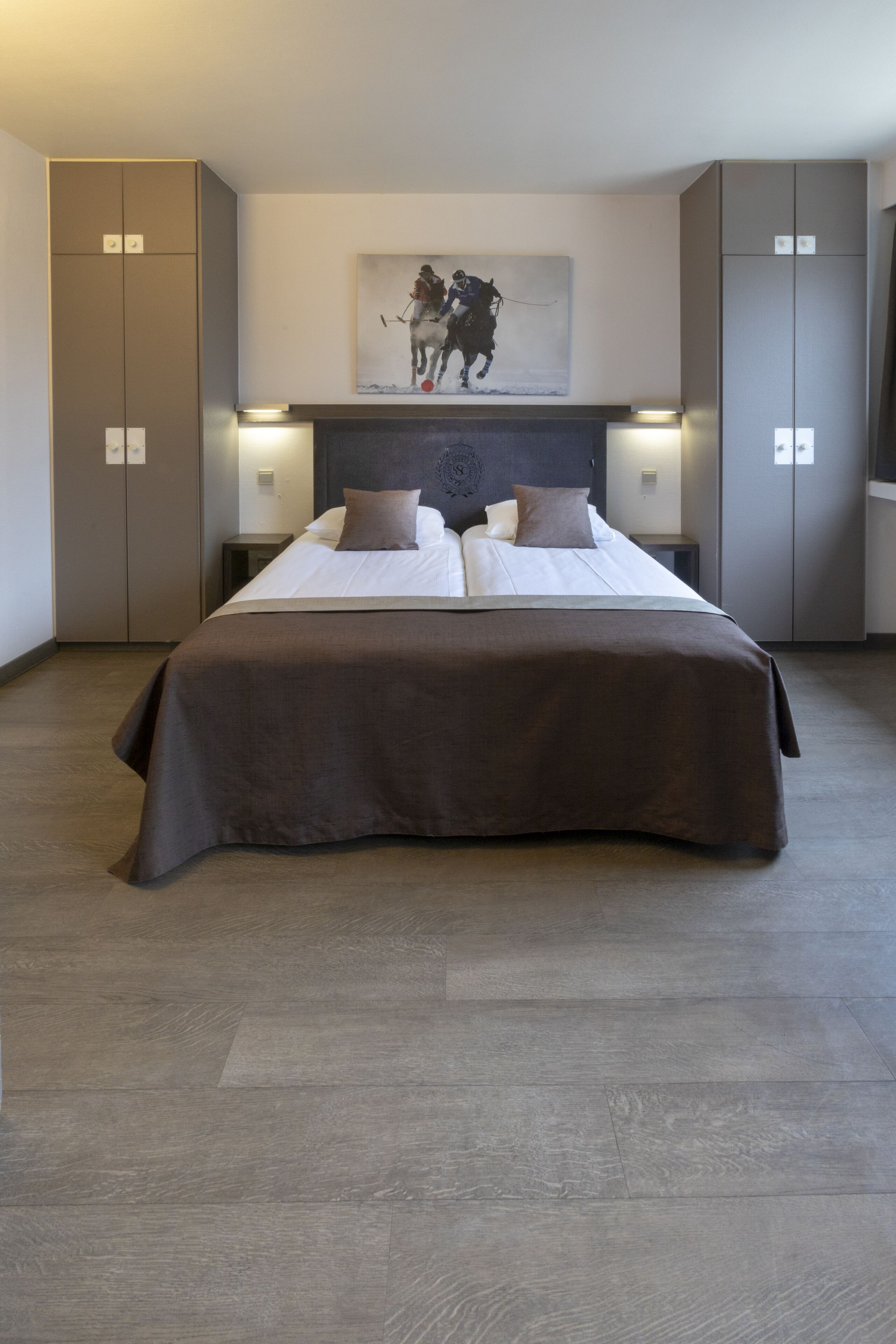 A modern bedroom with a large bed, two bedside tables, a headboard with a painting, and built-in wardrobes.