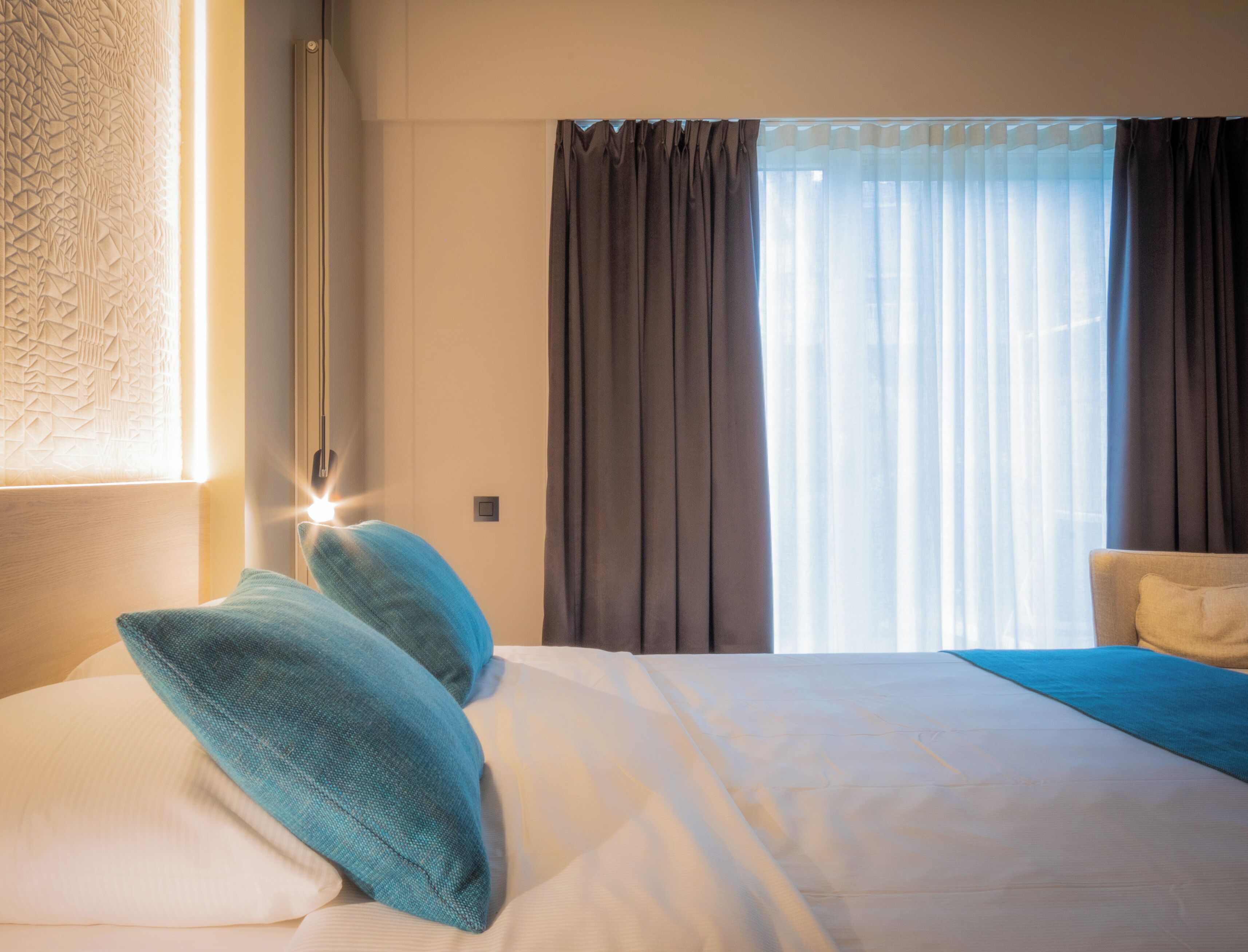 Una habitación de hotel con una cama bien hecha, una lámpara de noche y un ventanal con cortinas.