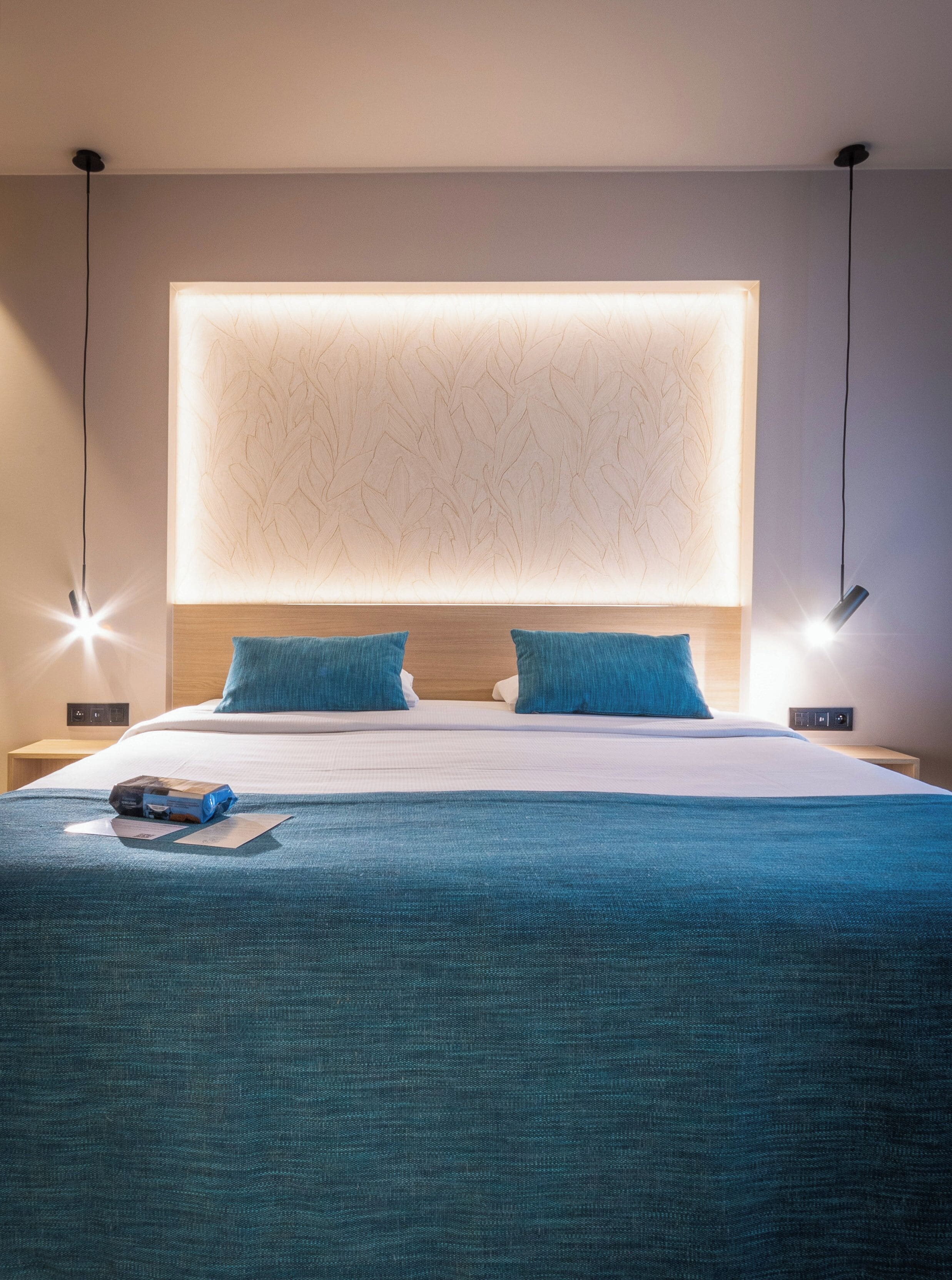 A modern hotel room with a large bed, two pillows, a bedside table with a lamp, and a wall-mounted light fixture.