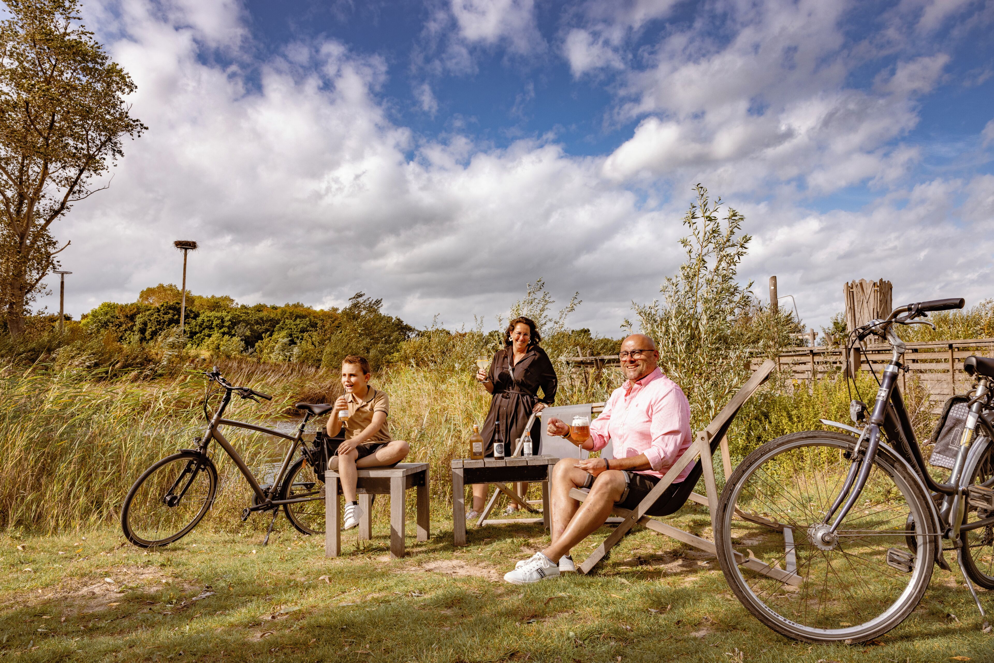 Trois personnes sont assises sur un banc, à côté de leurs vélos, dans un espace extérieur verdoyant, avec des arbres et un ciel nuageux.