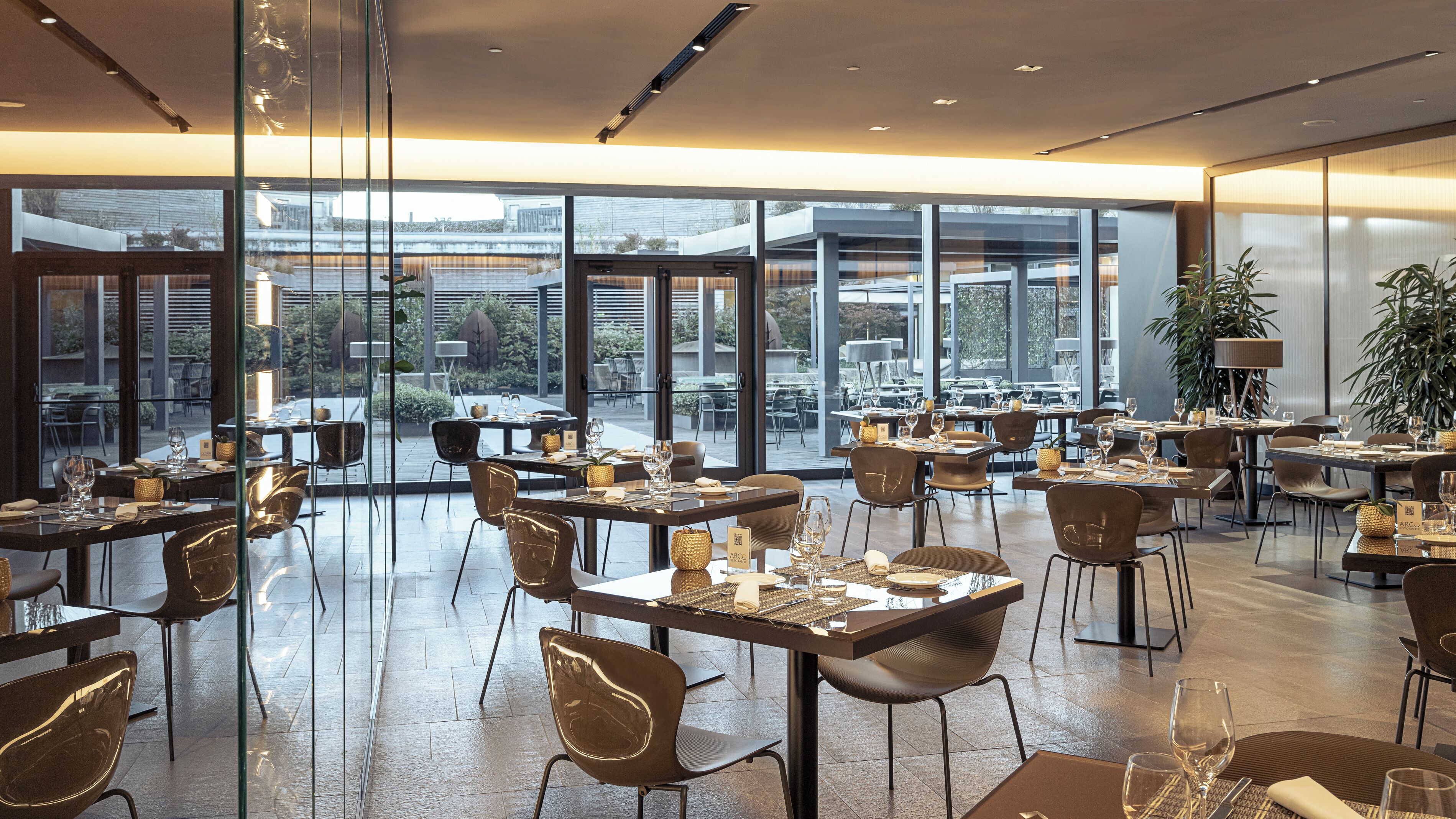 Ein modernes Restaurant mit mehreren Tischen und Stühlen, großen Fenstern mit Blick nach außen und einem klaren, zeitgemäßen Interieur.