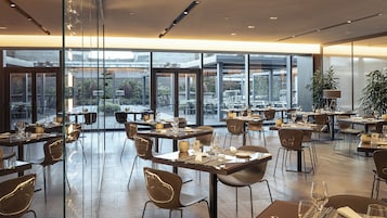 Un restaurante moderno con varias mesas y sillas, amplias ventanas que ofrecen vistas al exterior, y un diseño interior limpio y contemporáneo.