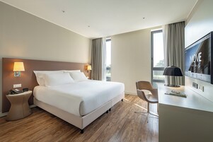 Habitación de hotel con cama, mesitas de noche, escritorio, silla, televisor y ventana con cortinas.