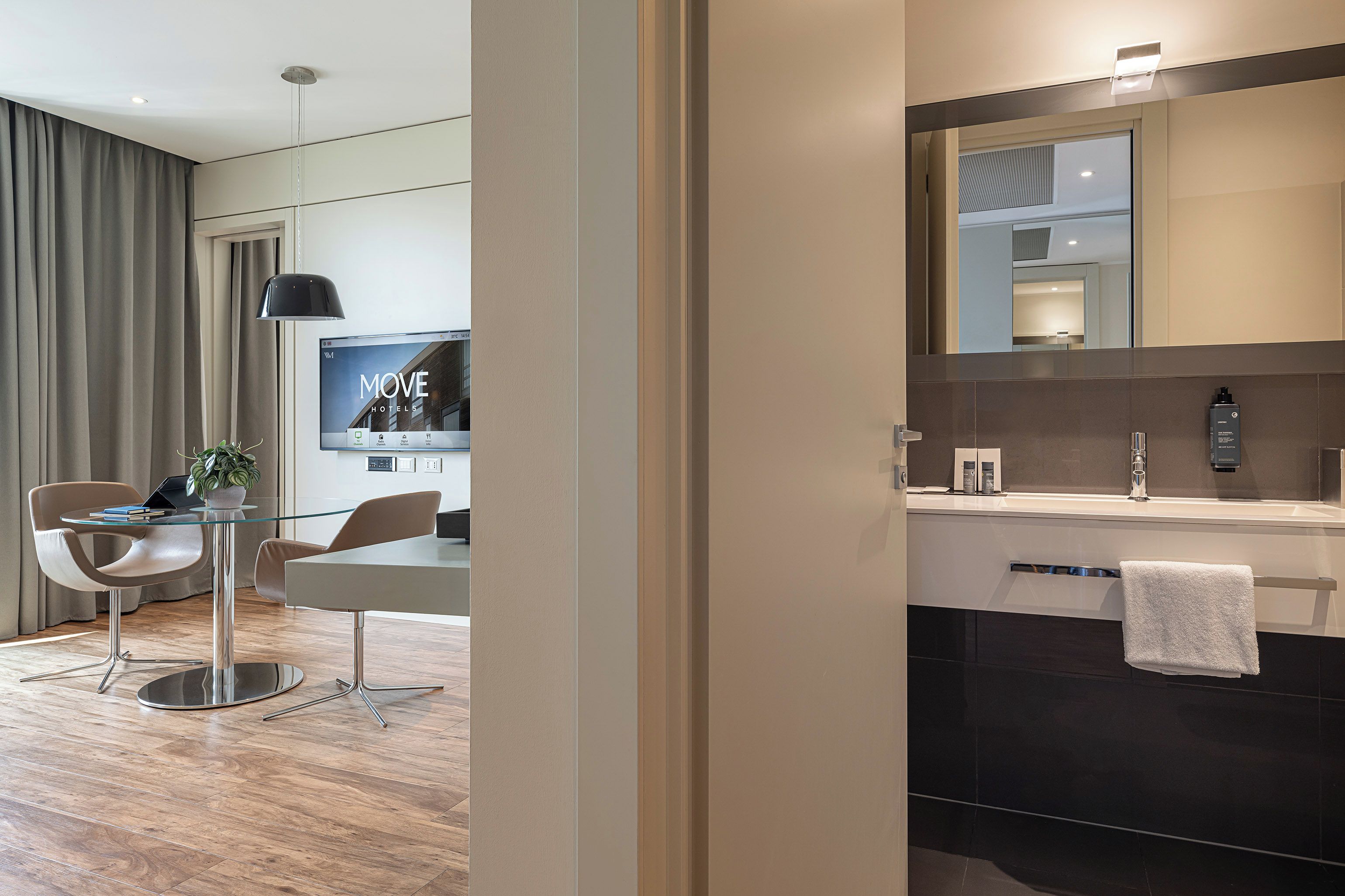 Quarto de hotel moderno com mesa de jantar de vidro, um laptop, uma planta em vaso e uma televisão exibindo 'MOVE HOTELS'.
