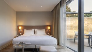 Quarto de hotel moderno com uma cama grande, abajures, uma mesa de vidro, um banco e vista para o exterior.