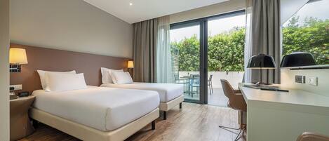A hotel room with two beds, a desk, and a view of greenery.