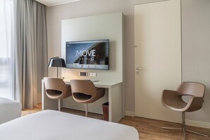 A hotel room with a bed, a desk with two chairs, a TV mounted on the wall, and a door.