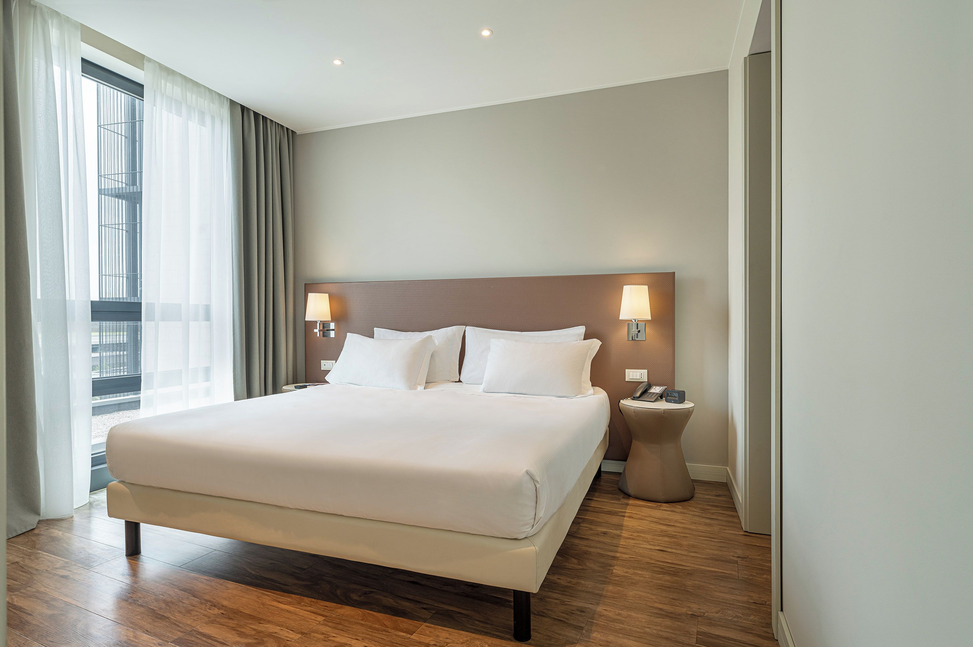 Habitación de hotel con una cama grande, dos lámparas de noche, una mesita de noche y vistas a la ciudad a través de la ventana.