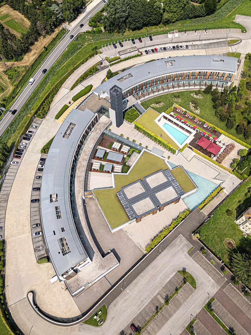 Vista aérea de um edifício moderno com telhado curvo, piscina e estacionamento.