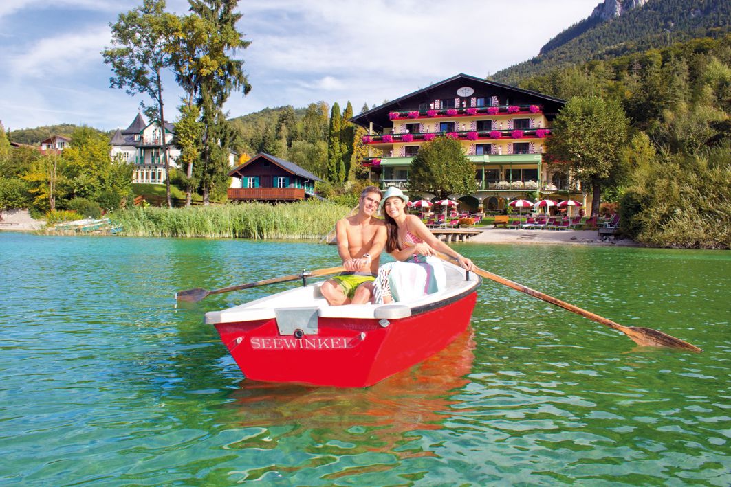 Un bote rojo con el nombre "SEEWINKEL" en un lago tranquilo rodeado de casas y árboles.