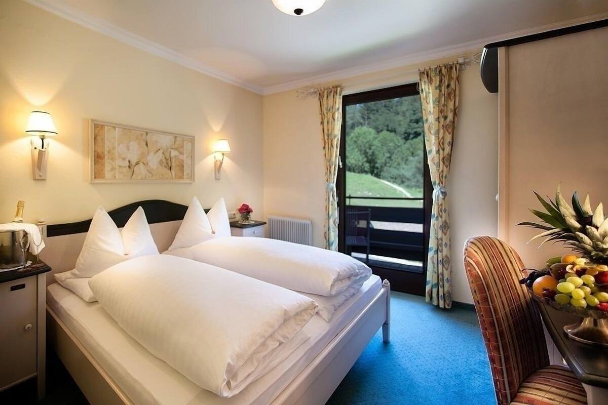 A hotel room with a double bed, two bedside lamps, a fruit bowl, a view of greenery through the window, and a painting on the wall.