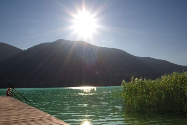 A wooden dock extending into a calm lake with a clear sky and mountains in the background.