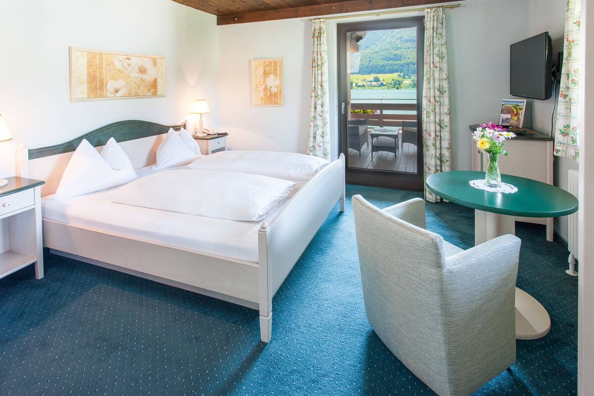 A hotel room with a bed, a small table with a vase of flowers, a chair, and a balcony with a view of the landscape.