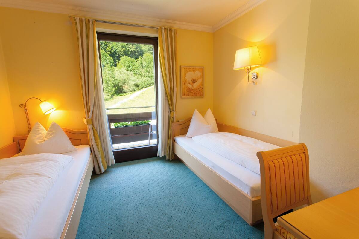 A hotel room with two single beds, a wooden chair, a table, a window with curtains, a wall-mounted lamp, and a painting.