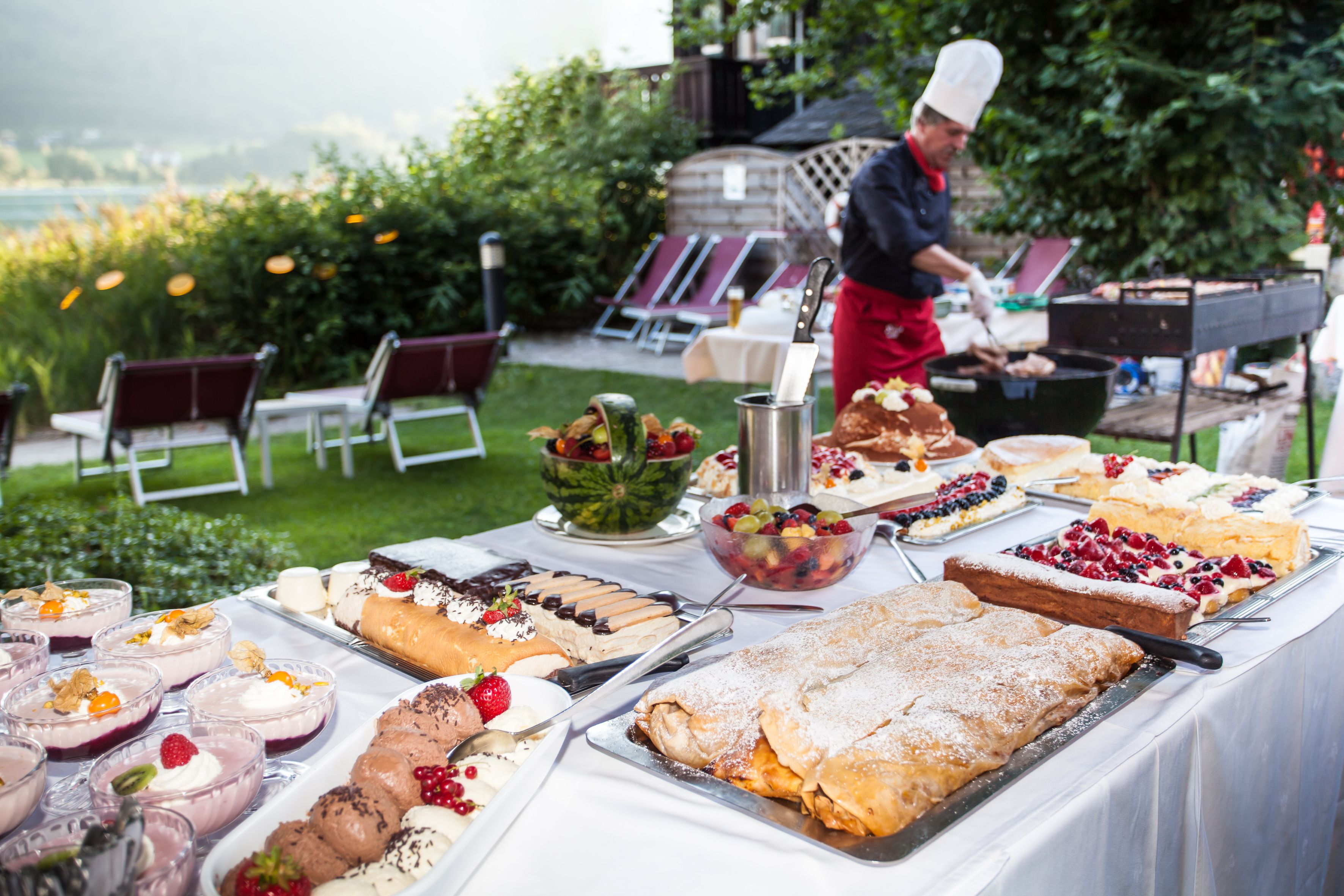 A buffet table with various desserts and a chef preparing food outdoors.