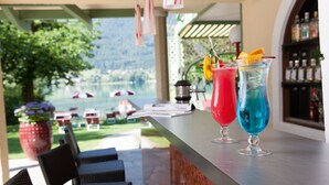 A bar area with two colorful drinks, outdoor seating, and a view of a lake. - Seewinkel (Fuschl am See)