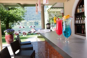 Zona de bar con dos bebidas coloridas, asientos al aire libre y vistas a un lago.