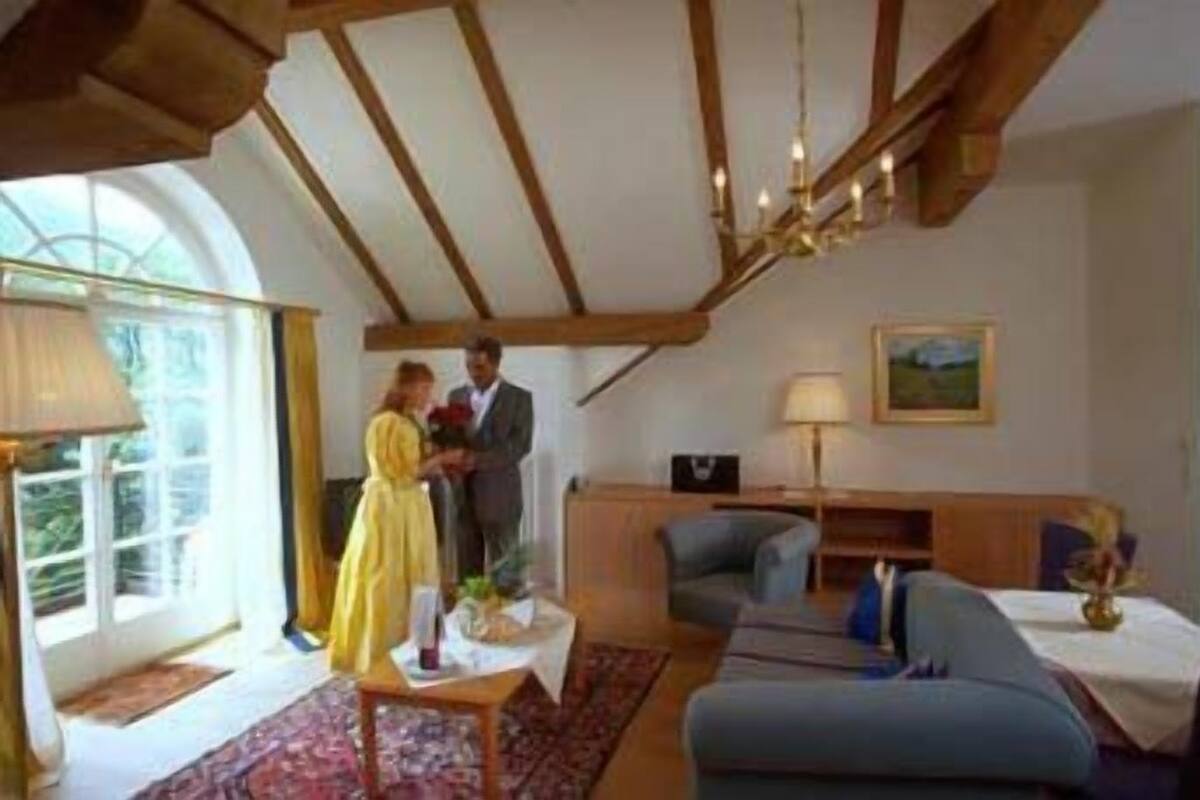 A room with a large window, a chandelier, a sofa, a chair, and a table with a lamp.