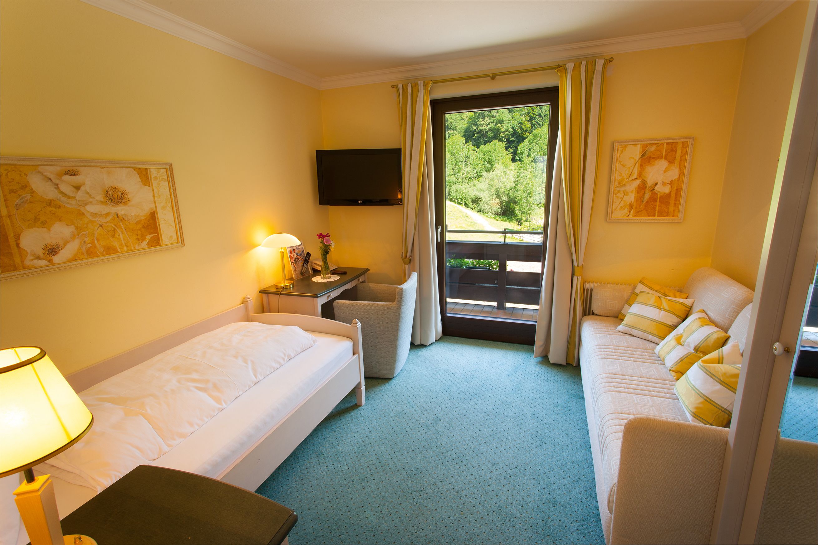 Ein Hotelzimmer mit Bett, Sofa, Schreibtisch mit Lampe, Fernseher und Blick auf Grünanlagen.