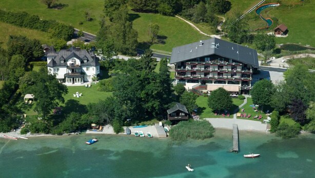 An aerial view of a coastal hotel with multiple buildings, including a large hotel with balconies and a smaller lodge. There are boats docked near the shore, and the area is surrounded by greenery. - Seewinkel (Fuschl am See)