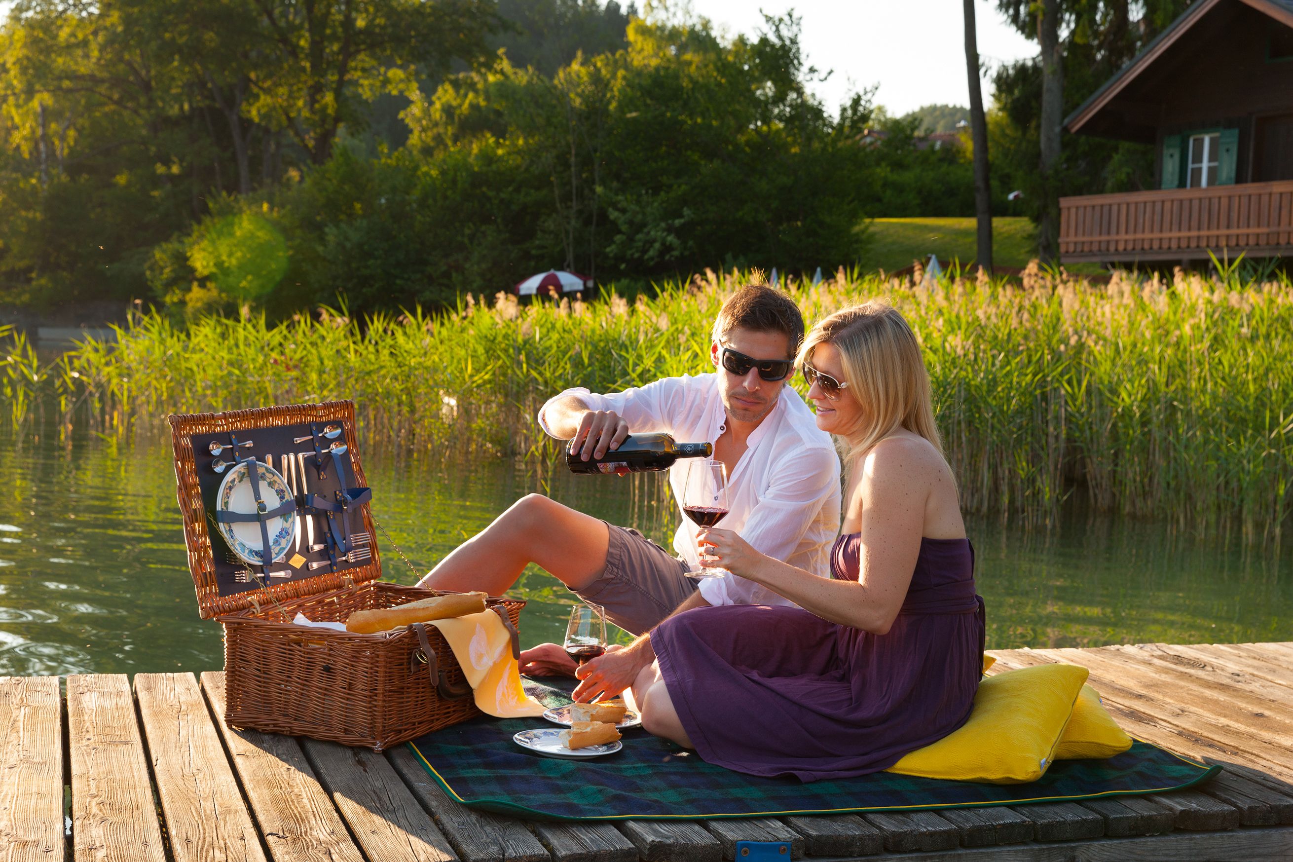 A wooden dock with a picnic setup including a basket, wine glasses, and a blanket.