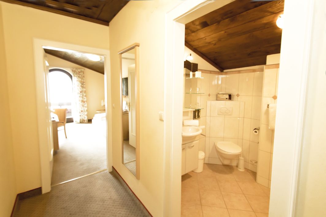 Pasillo que conduce a un baño con inodoro, lavamanos y espejo. El techo es de madera y hay una puerta que da a otra habitación.