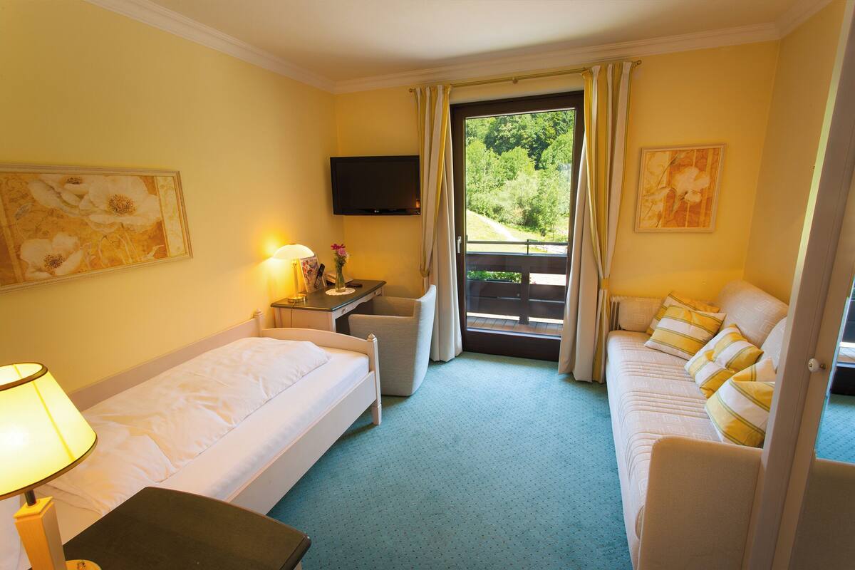 A hotel room with a bed, a sofa, a desk with a lamp, a TV, and a window with curtains.