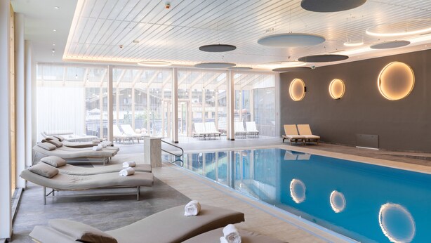 Una piscina interior moderna con sillones de descanso y tumbonas, ventanas amplias y lámparas circulares en el techo.