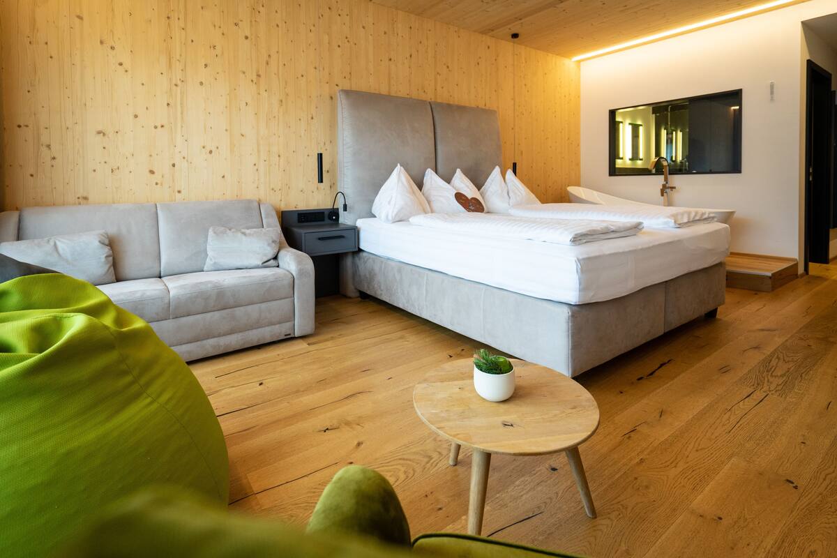 A modern hotel room with a wooden floor, a grey sofa, a bed with white linens, a small round table with a plant, and a wooden headboard.