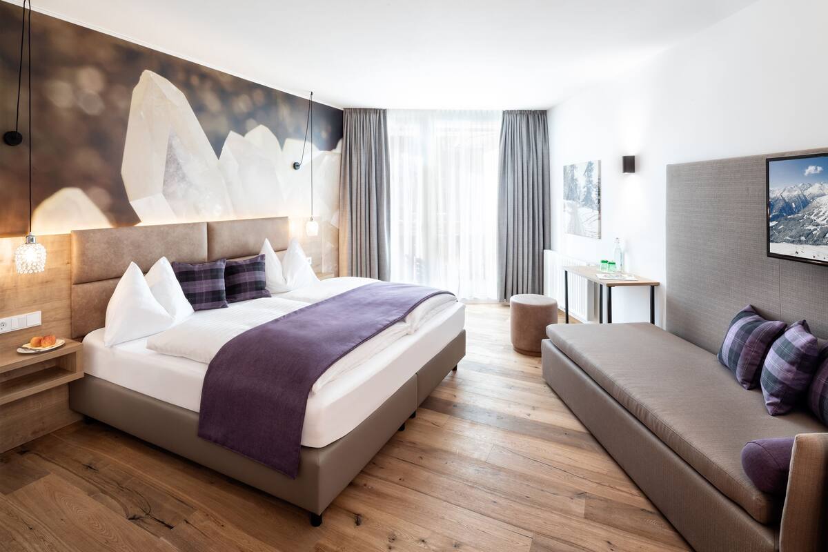 A modern hotel room with a large bed, a sofa, wooden flooring, a wall-mounted TV, and a window with curtains.