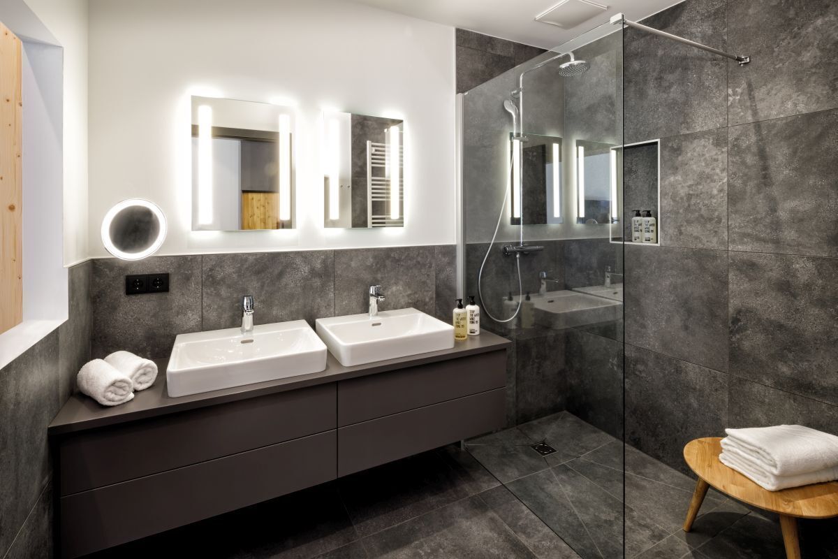 A modern bathroom with two sinks, a glass shower enclosure, and a wooden stool with towels.