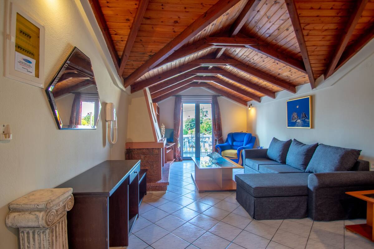 Ein großzügiger Wohnbereich mit hoher Holzbalkendecke, einer Couch, einem Couchtisch und Blick durch eine Glastür auf einen Außenbalkon.