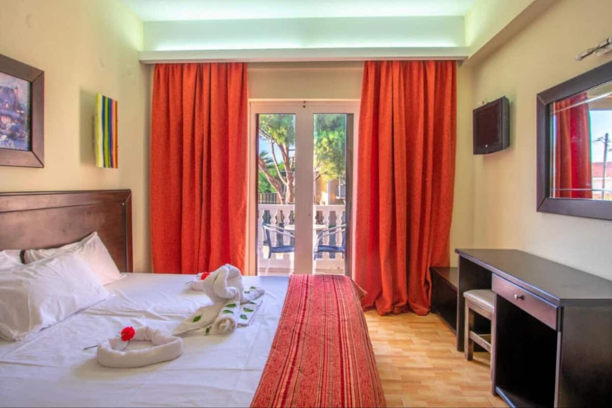 Ein Hotelzimmer mit Bett, Schreibtisch, Fernseher und einem Balkon mit roten Vorhängen.