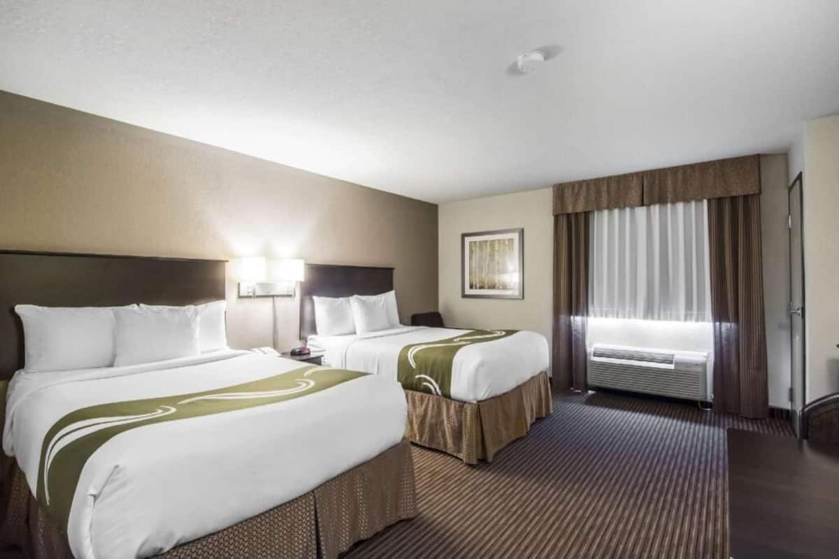 A hotel room with two beds, a large window with curtains, a wall-mounted air conditioner, and a framed picture on the wall.